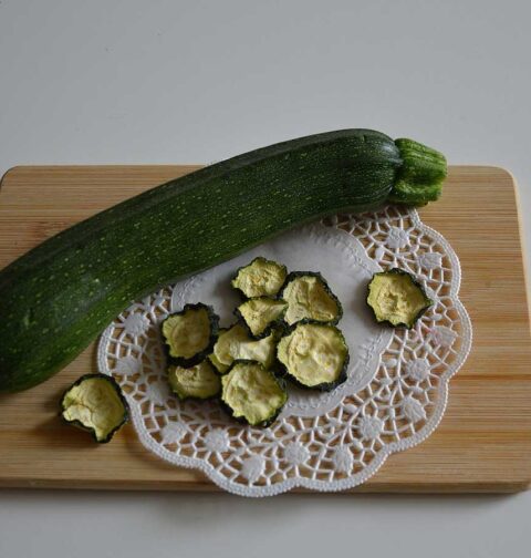 Gedroogde plakjes courgette naast een hele courgette op een houten snijplank.