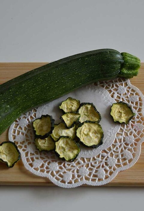 Gedroogde plakjes courgette naast een hele courgette op een houten snijplank.
