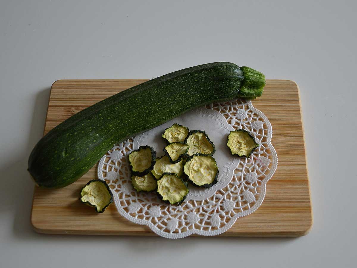 Gedroogde plakjes courgette naast een hele courgette op een houten snijplank.