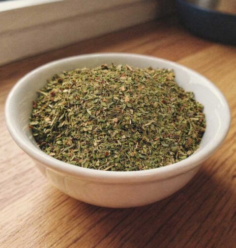 Een wit keramisch kommetje gevuld met fijngehakte gedroogde oregano op een houten aanrecht.