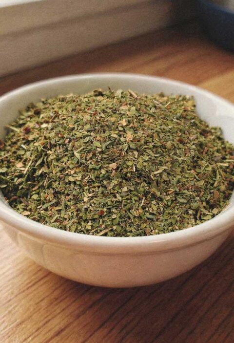 Een wit keramisch kommetje gevuld met fijngehakte gedroogde oregano op een houten aanrecht.