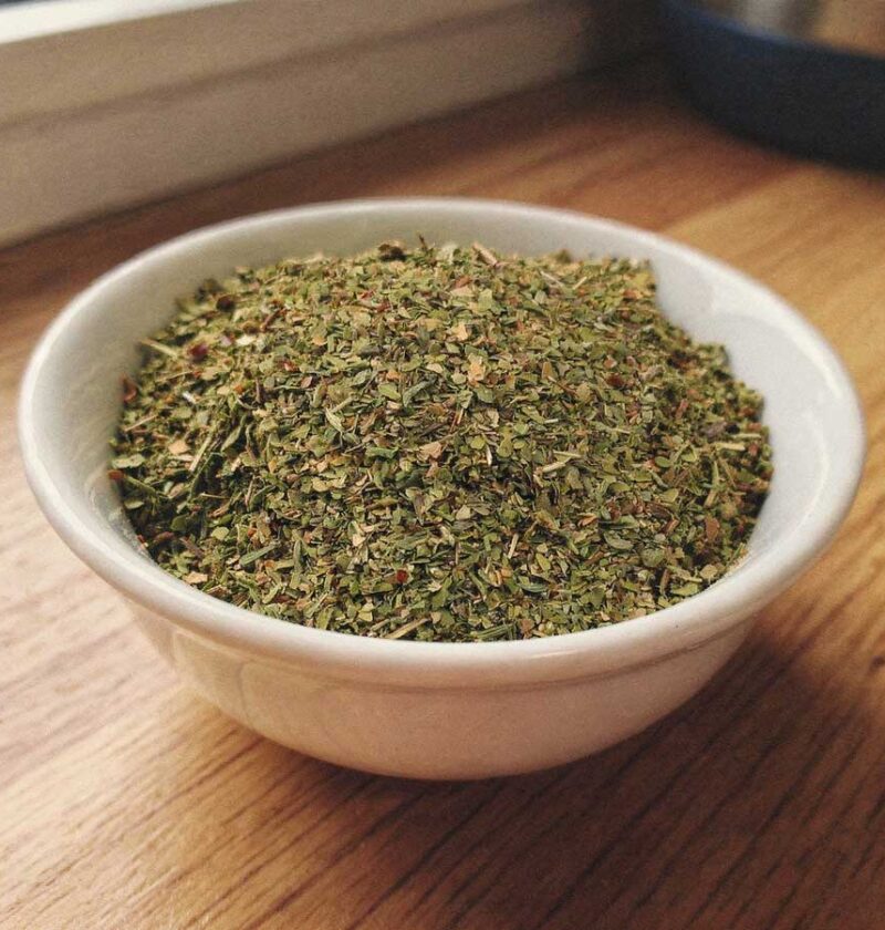 Een wit keramisch kommetje gevuld met fijngehakte gedroogde oregano op een houten aanrecht.