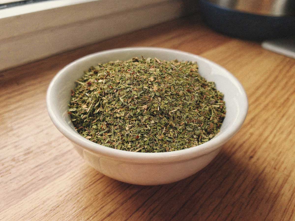 Een wit keramisch kommetje gevuld met fijngehakte gedroogde oregano op een houten aanrecht.