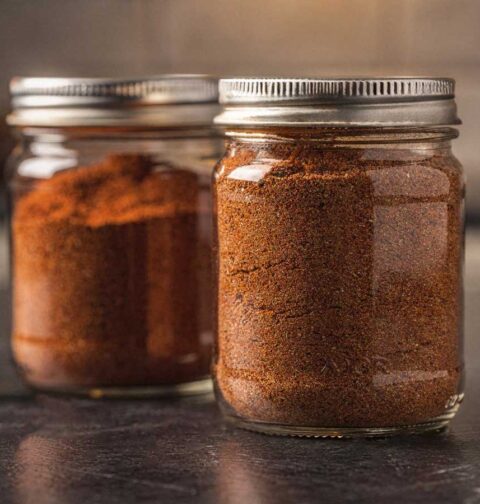 Twee glazen potjes gevuld met zelfgemaakte goudbruine BBQ rub op een donker aanrecht.