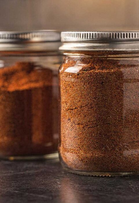 Twee glazen potjes gevuld met zelfgemaakte goudbruine BBQ rub op een donker aanrecht.
