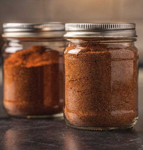 Twee glazen potjes gevuld met zelfgemaakte goudbruine BBQ rub op een donker aanrecht.