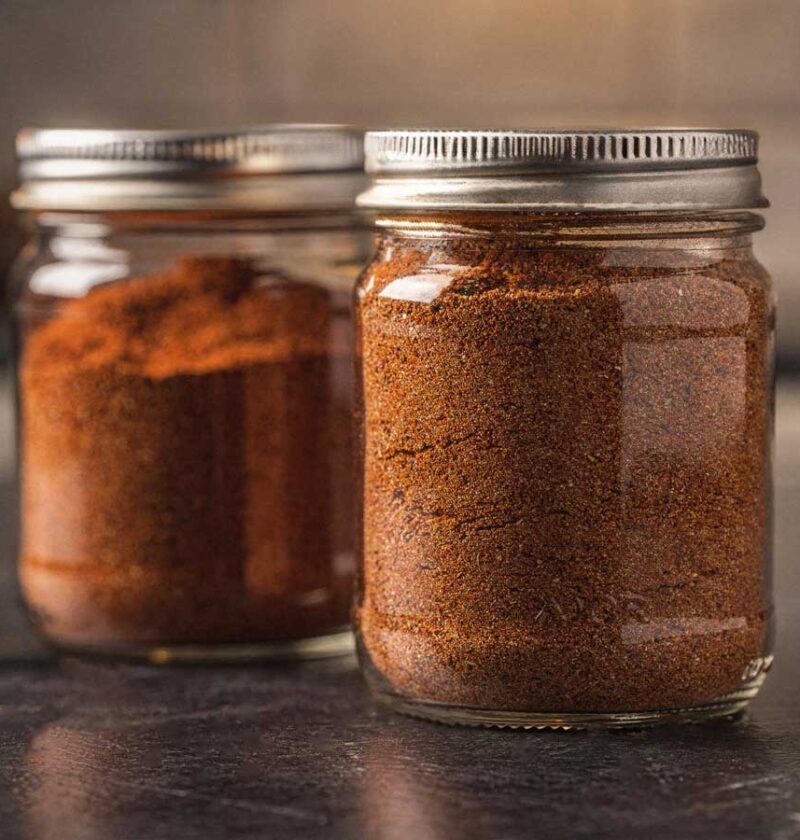 Twee glazen potjes gevuld met zelfgemaakte goudbruine BBQ rub op een donker aanrecht.