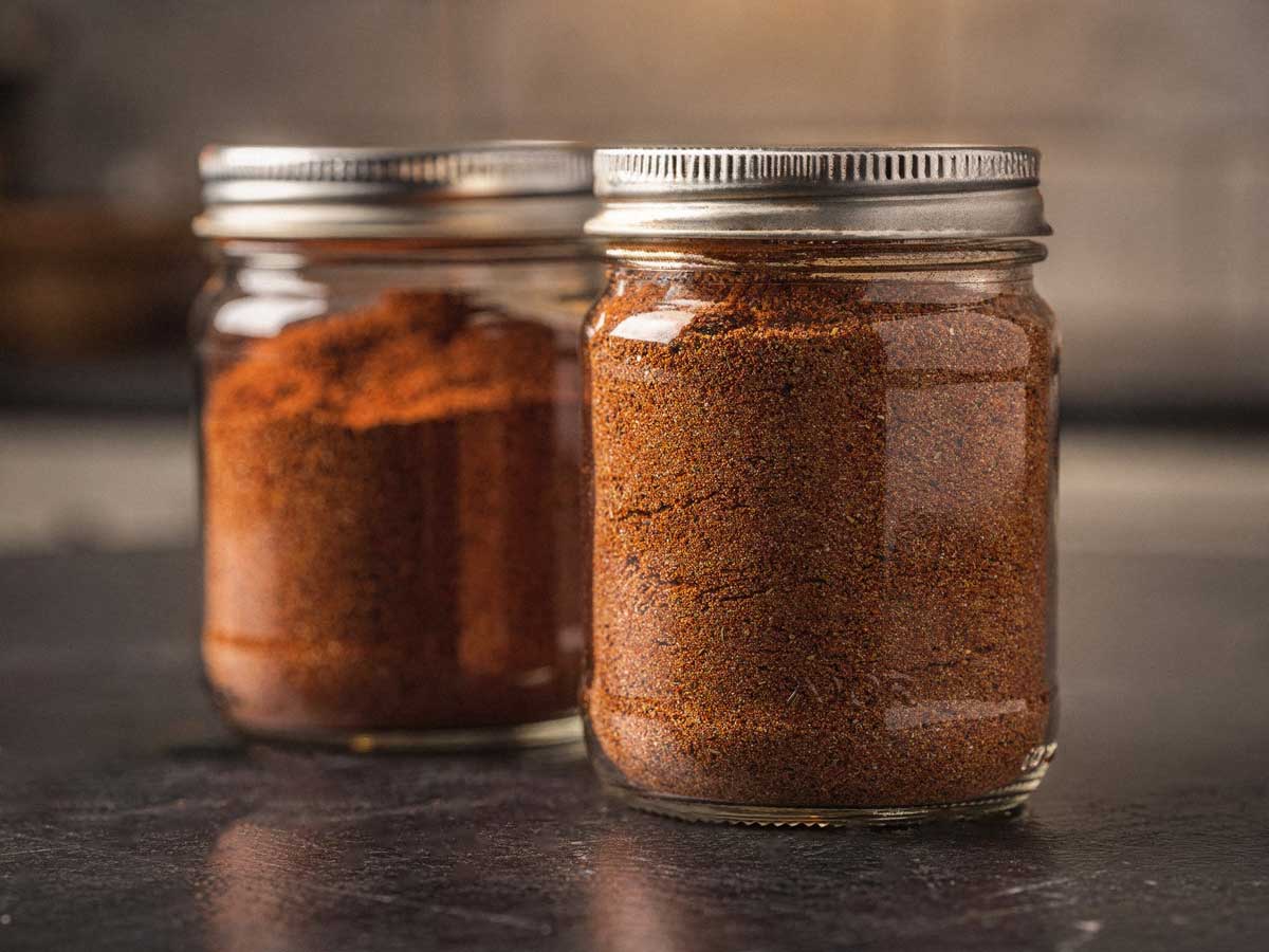 Twee glazen potjes gevuld met zelfgemaakte goudbruine BBQ rub op een donker aanrecht.