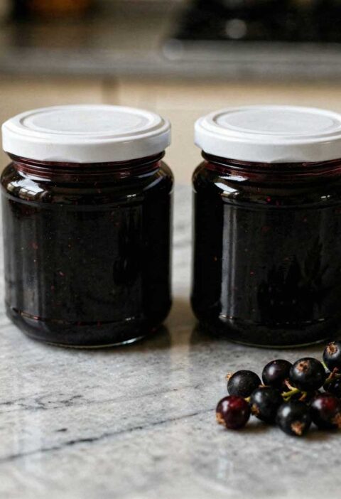 Twee glazen jampotten met witte deksels gevuld met zwarte bessenjam op een marmeren aanrecht